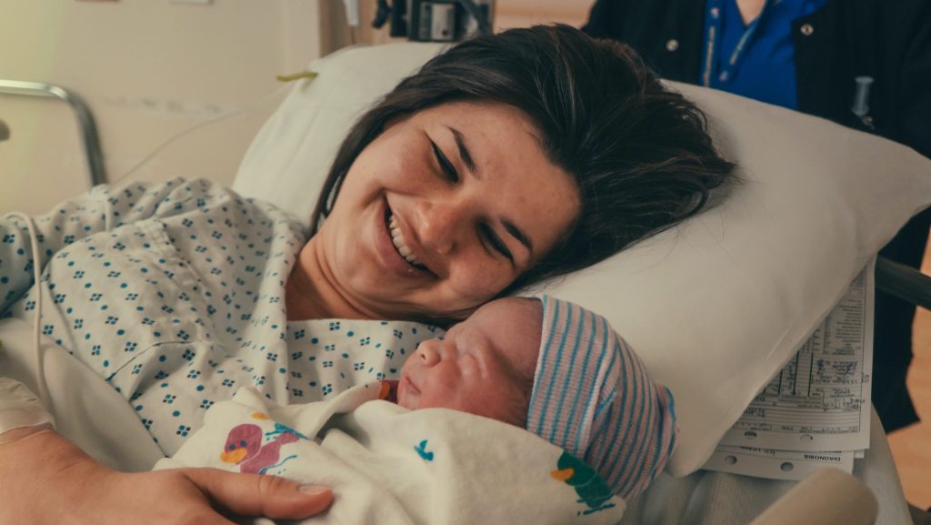 A smiling woman in a hospital bed holds a newborn wrapped in a white blanket with colorful patterns. The scene conveys warmth and joy.
