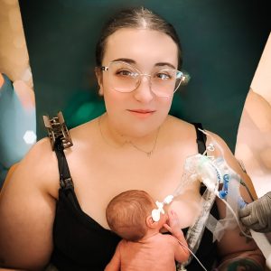 A mom wearing a tank top sits in a chair holding her newborn son who is sleeping on her chest.