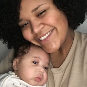 Smiling person with curly hair holding a resting baby in a cozy embrace. Both wear light-colored clothing, creating a warm, tender atmosphere.