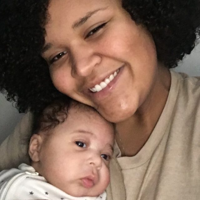 Smiling person with curly hair holding a resting baby in a cozy embrace. Both wear light-colored clothing, creating a warm, tender atmosphere.