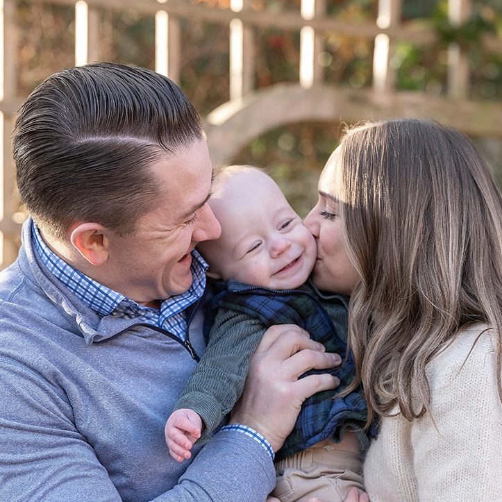 A joyful family scene with a man holding a baby, who is smiling as a woman kisses the baby’s cheek. They stand outdoors, surrounded by greenery.